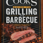Book cover of Cook's Illustrated Guide to Grilling and Barbecue featuring a thick, perfectly grilled steak sliced on a hot barbecue grate above glowing flames. The black cover design highlights bold white lettering and promises expert barbecue techniques and recipes.
