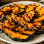 A rustic ceramic plate filled with grilled acorn squash slices sits on a weathered wooden table. The squash features deep caramelized grill marks and golden-orange flesh framed by dark green skin. Fresh herbs are sprinkled over the top, adding vibrant green accents. The squash appears tender and lightly charred, showcasing a sweet and smoky finish.