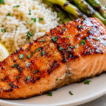 Grilled glazed salmon fillet with herb rice, charred asparagus, and a lemon wedge on a white plate.